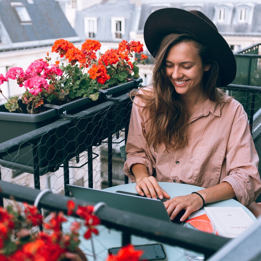 woman wearing a hat surrounded by flowers looking at laptop